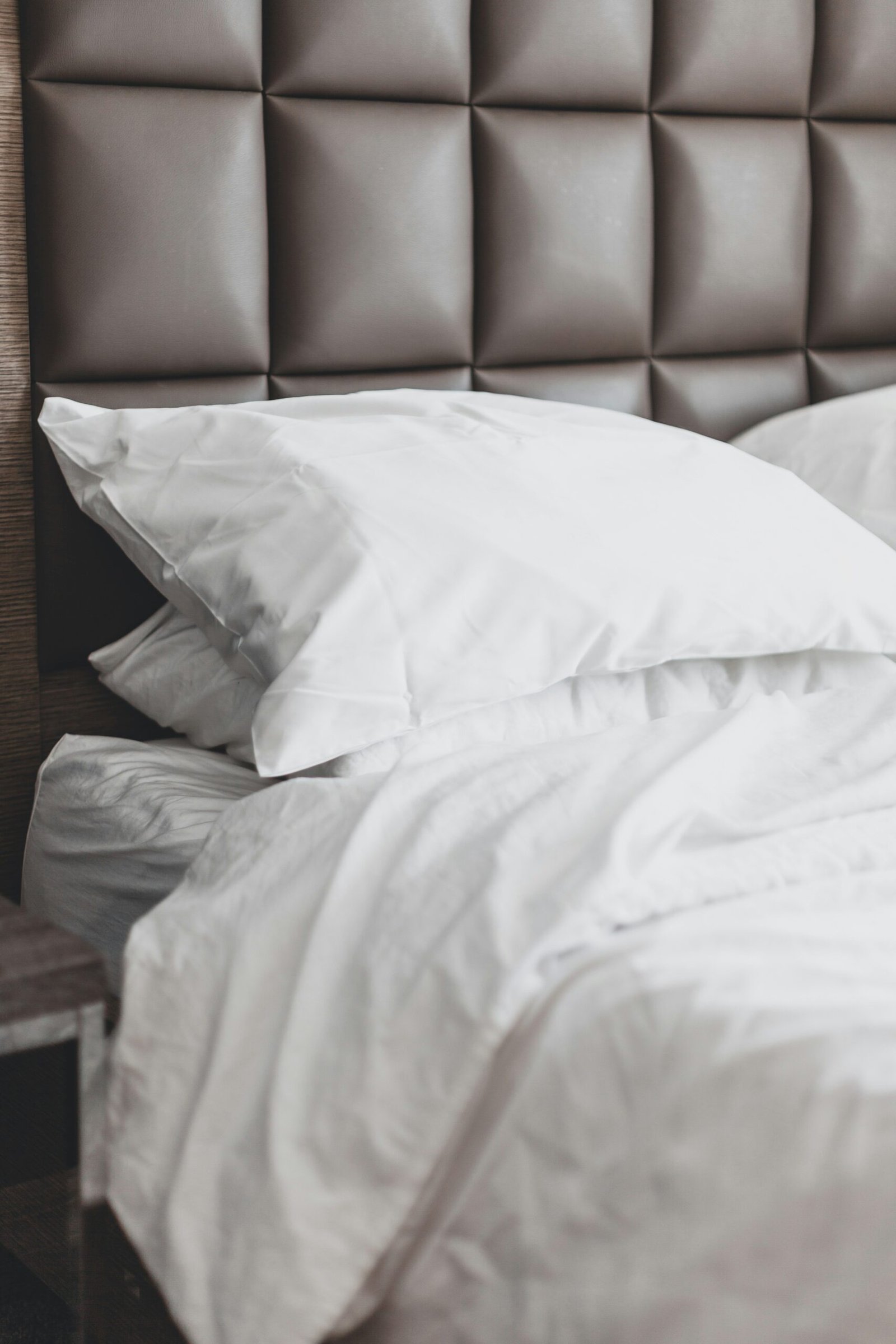 Close-up of a bed with white pillows and rumpled white bedding against a padded brown headboard.

